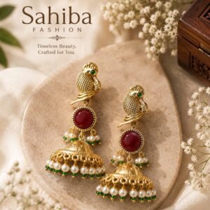 Pair of Sahiba Fashion golden peacock jhumka earrings featuring red cabochon stones and pearl drops on a stone slab.